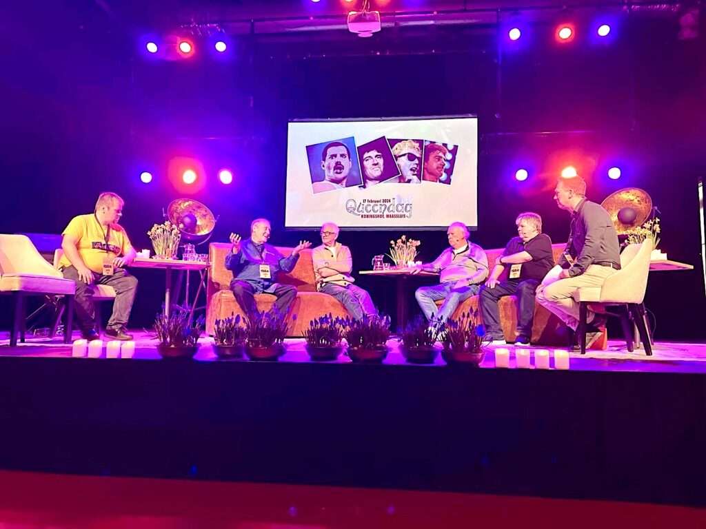 Groep mannen op een podium, zittend op stoelen en banken. Ze lijken een gesprek te voeren. Op de achtergrond een foto van de 4 bandleden van Queen, met daaronder de tekst 17 februari 2024 Queendag - Koningshof, Maassluis