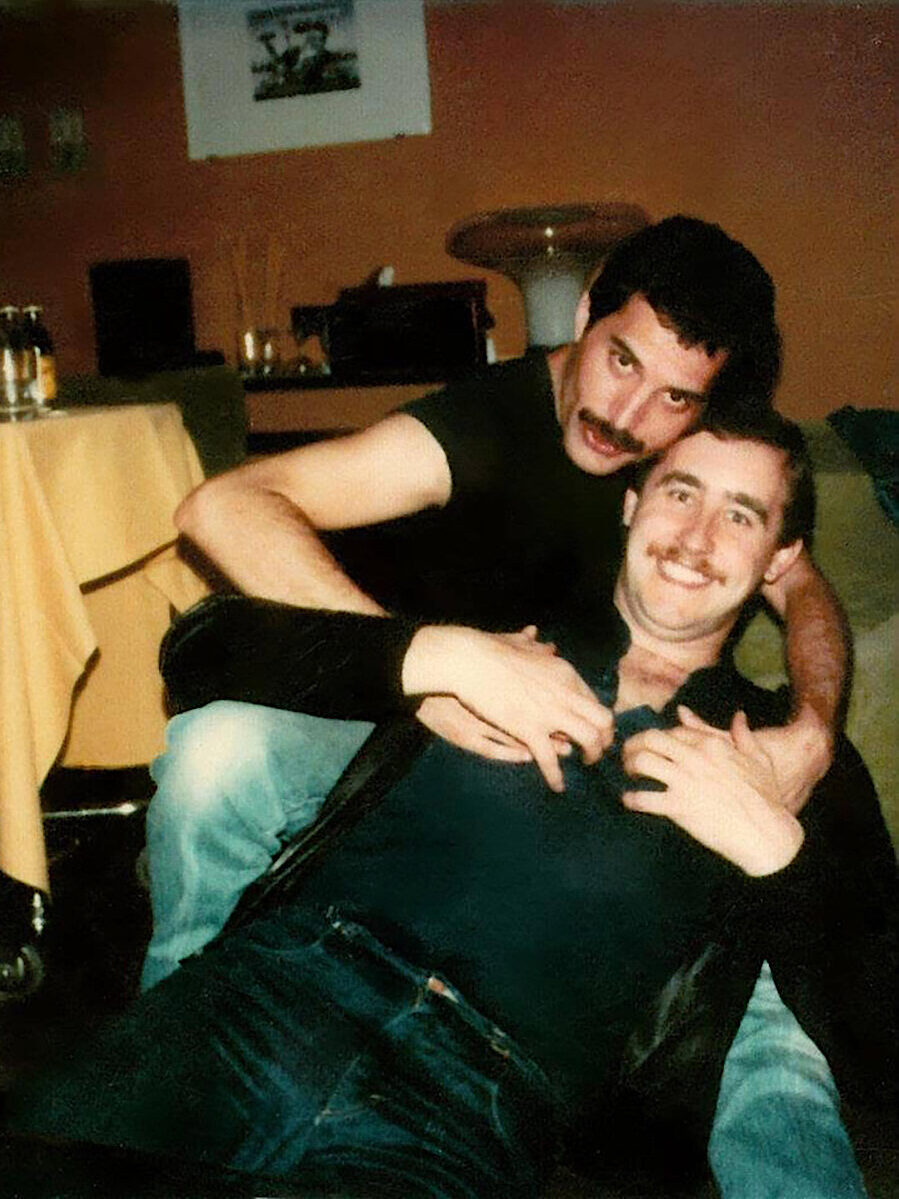 2 mannen, zittend achter elkaar, achterste man houdt zijn armen om de voorste en de voorste heeft de handen van de achterste vast.
Man op de achtergrond is Freddy Mercury en op de voorgrond is Peter Freestone.
