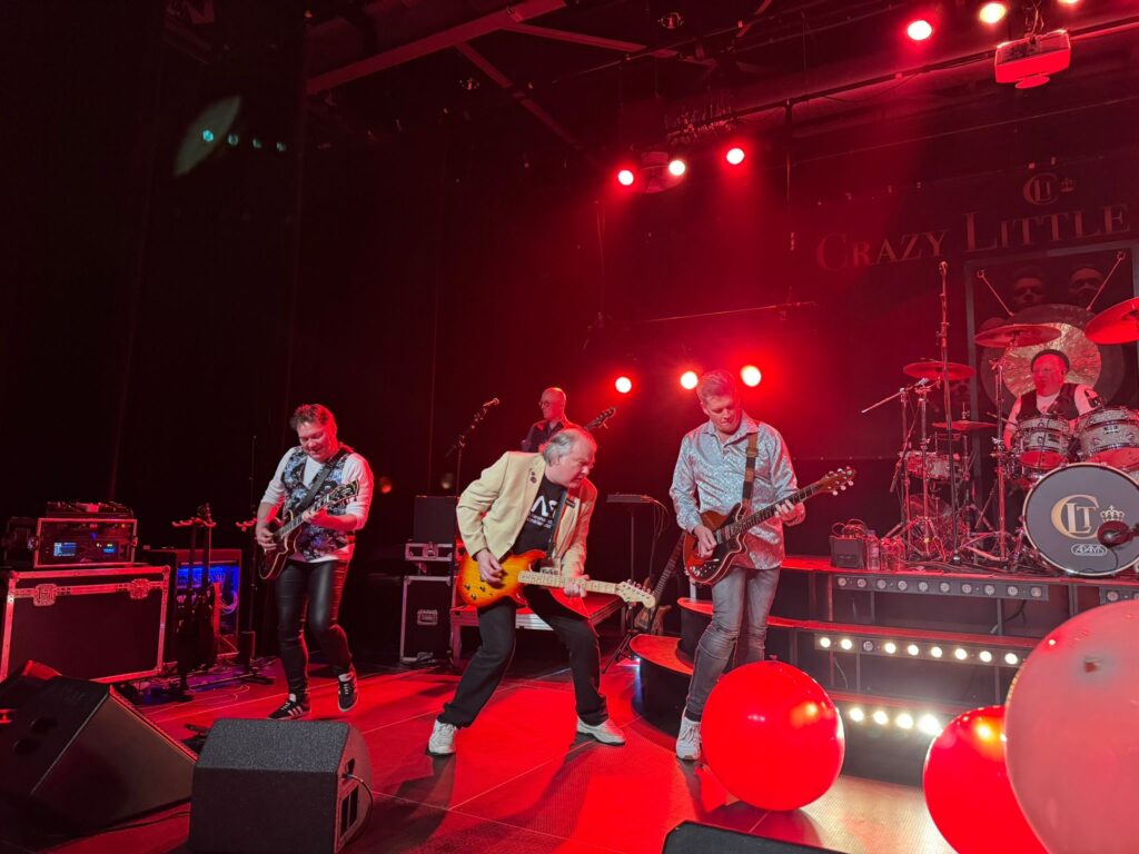 Band op een podium, verlicht door rode lampen. 3 gitaarspelers, op de achtergrond een basspeler en een drummer.