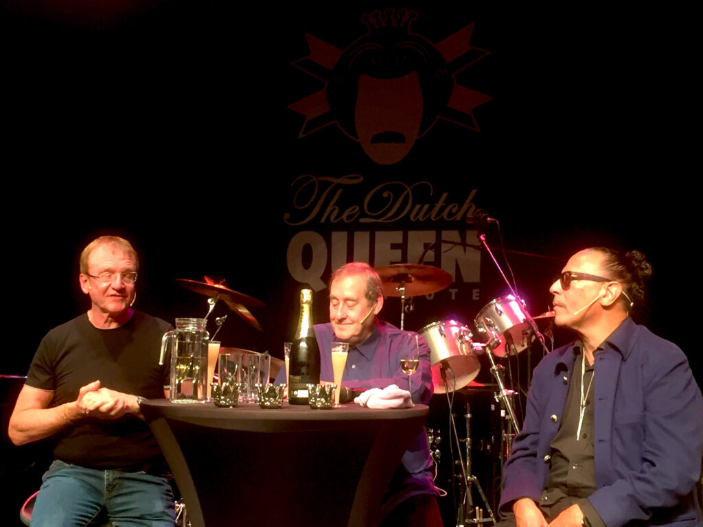 3 mannen achter een tafeltje waarop glazen, een waterkan en een fles champagne staan. Achter de mannen is half te lezen 'The Dutch Queen...' te lezen en er staat een drumstel.